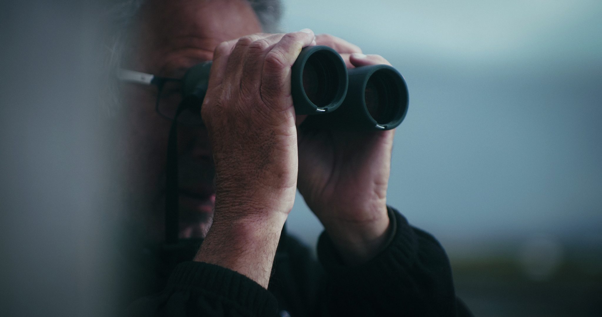 A man with greying hair holds binoculars up to his eyes, intently observing the distant landscape on a cloudy day.