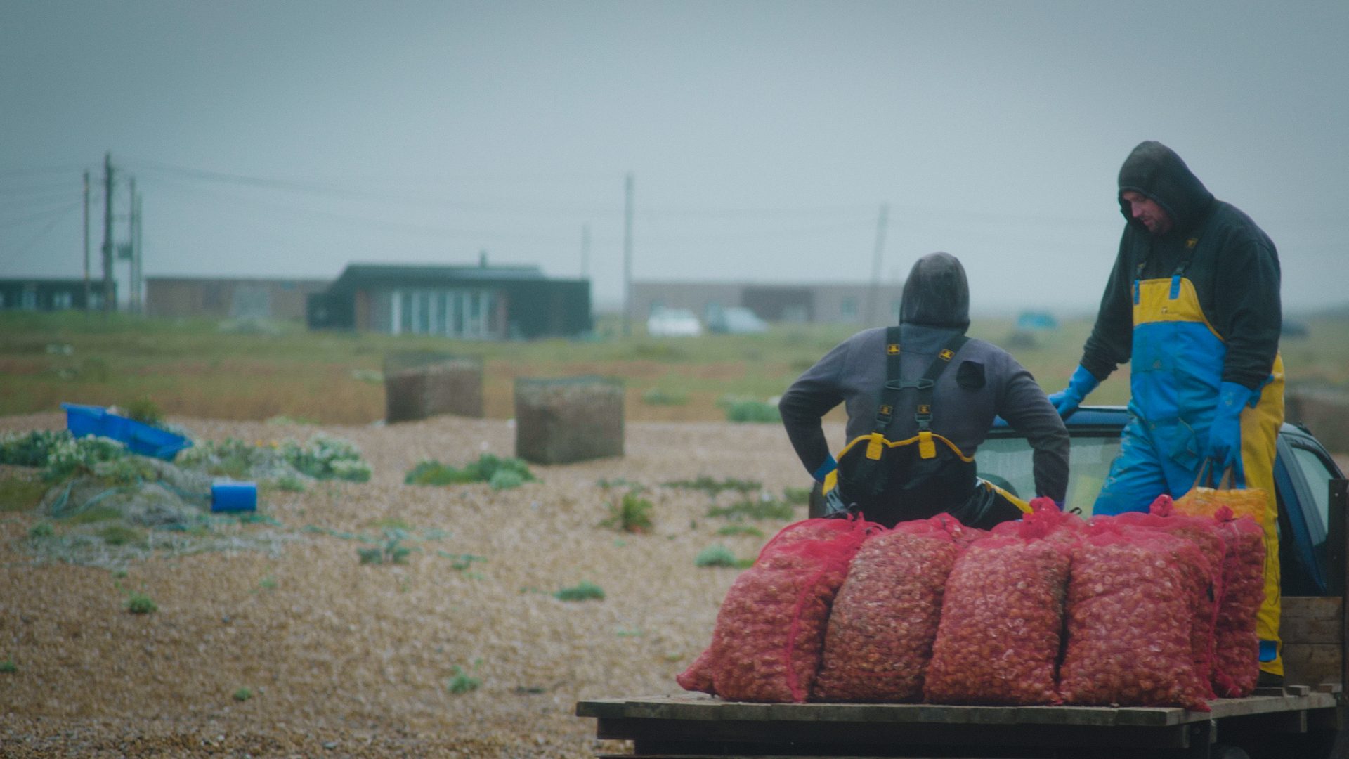 Two fishermen in rain gear stand near bags of freshly caught seafood and a vehicle on a misty, pebbled beach.