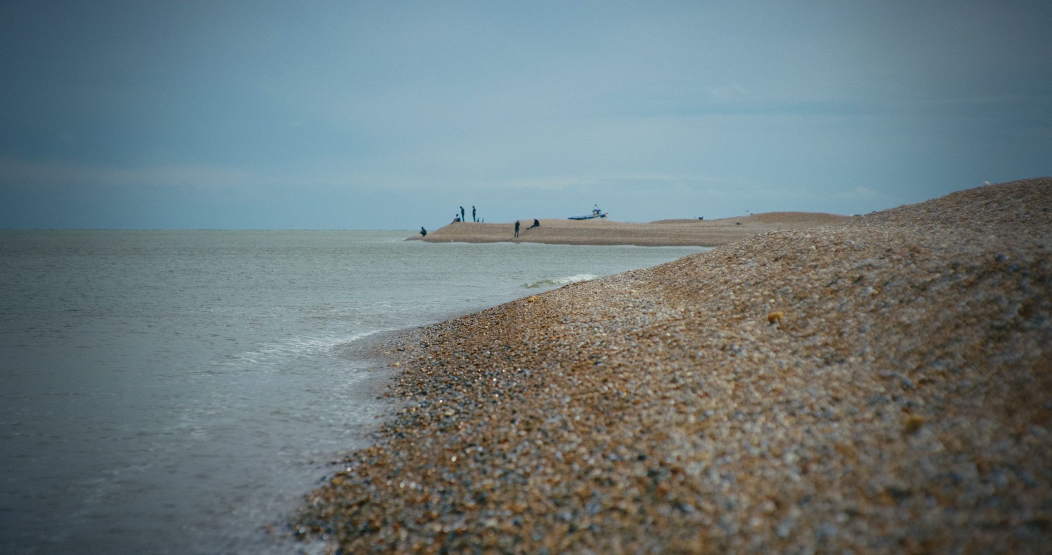 A shoreline scene featuring a pebbly beach extending into the calm sea, with figures silhouetted against a cloudy blue sky.