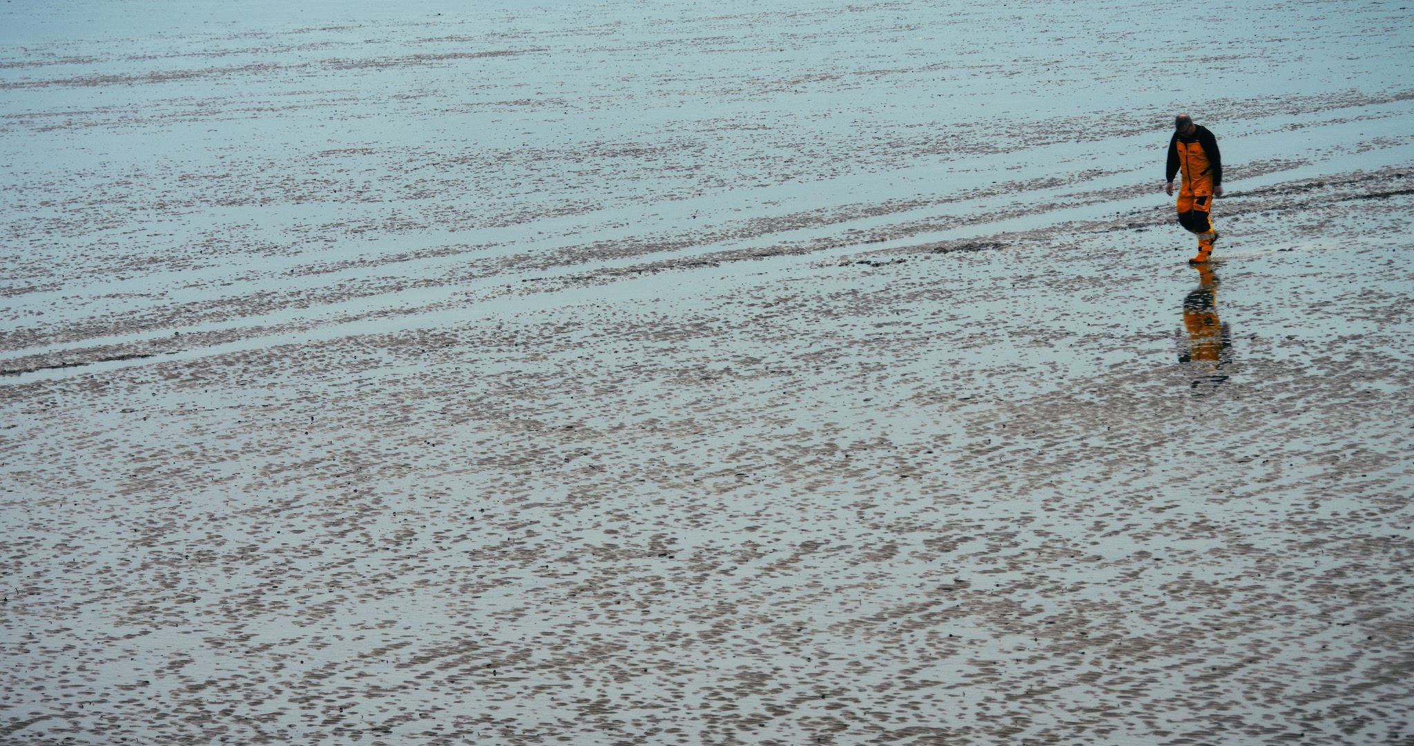 A solitary lifeboatman in a bright orange outfit walks along a wet, reflective beach, leaving footprints in the sand.