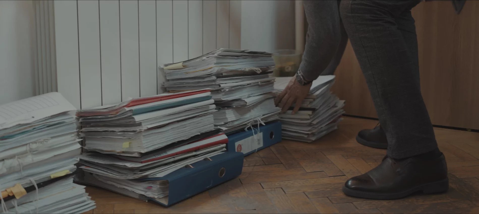 A man in a suit bends to sort stacks of case files and binders piled on a parquet floor.